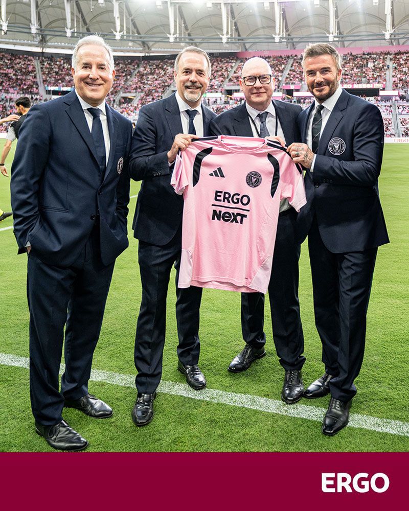 Four men in suits are standing on a football pitch, holding up an Inter Miami shirt for the camera.