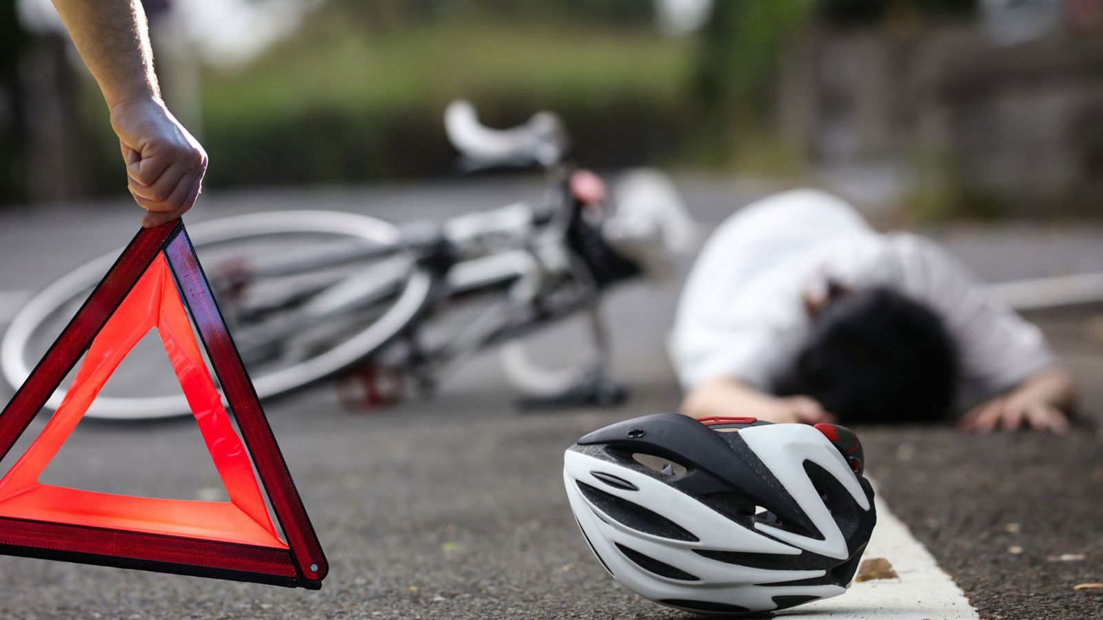 A person is lying on the ground next to a bicycle and a helmet, with a warning triangle visible in the foreground.