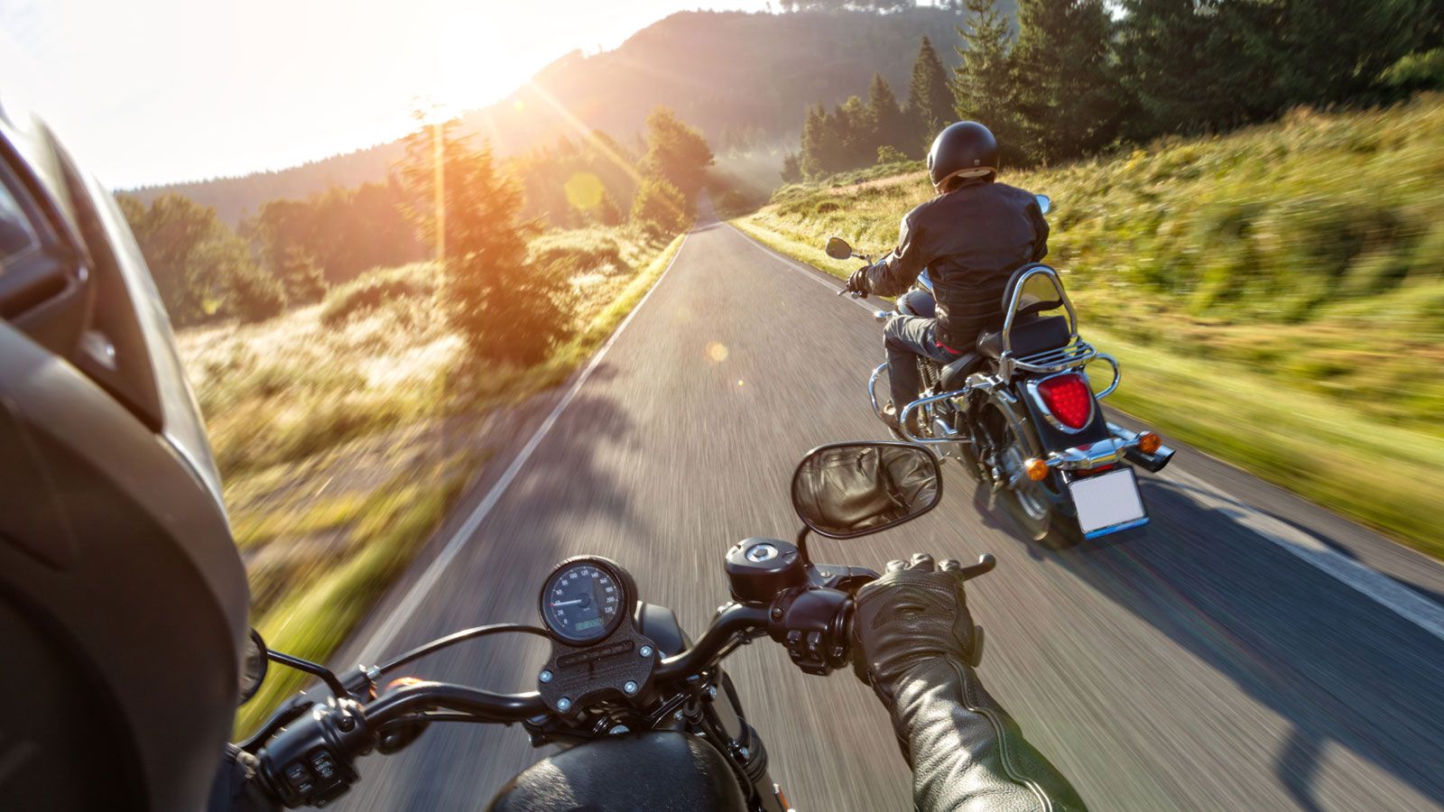 A view over the shoulder of a motorcyclist looking down a country road with another motorcycle ahead at sunset