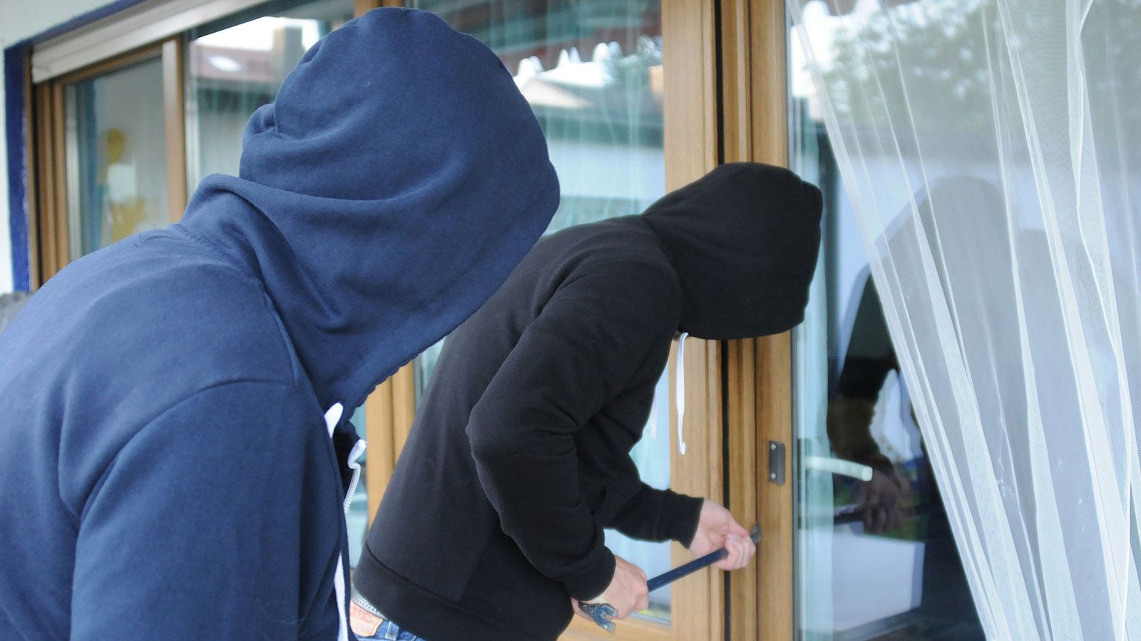 Two people wearing hoodies are trying to pry open a window with a crowbar