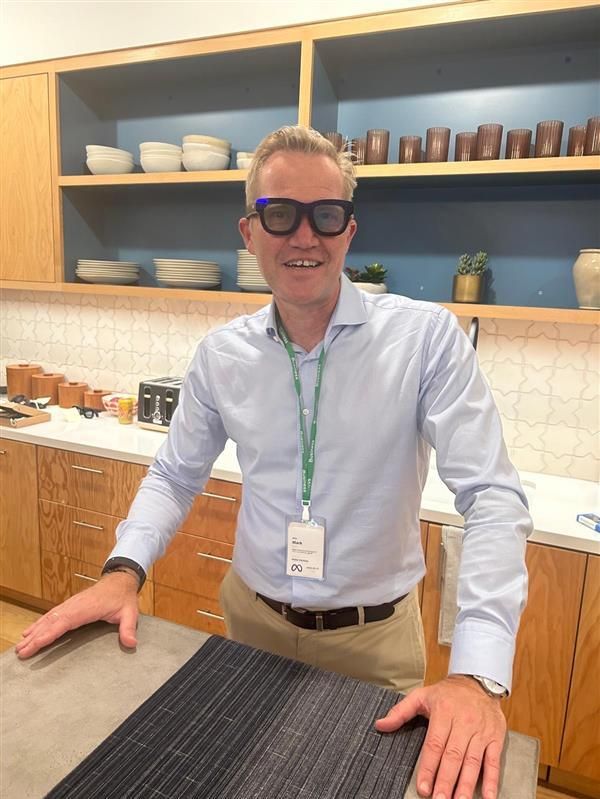 A man is standing in a kitchen wearing distinctive glasses.