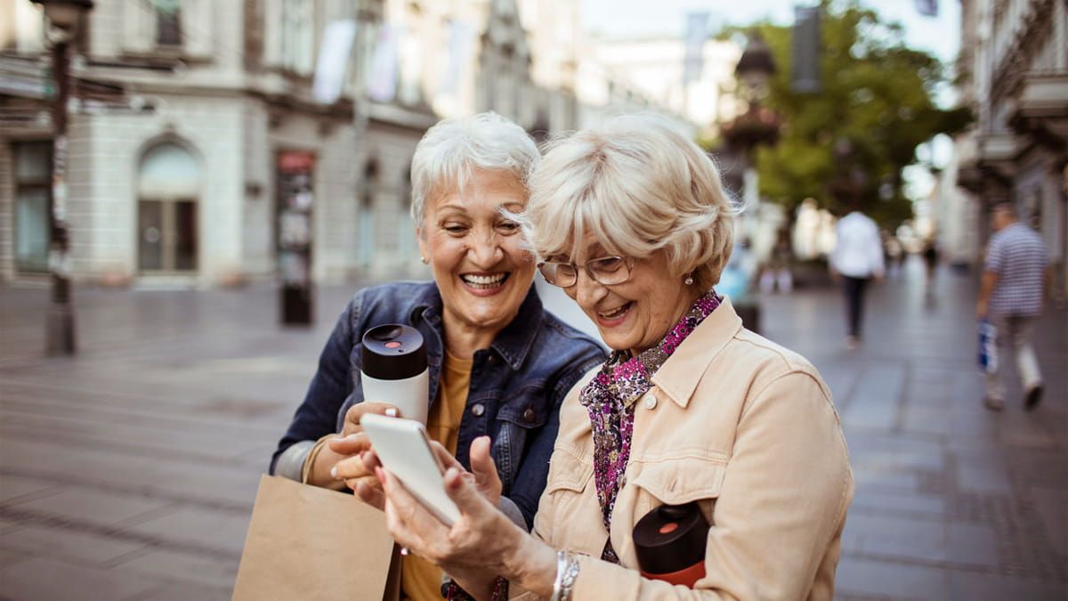 Zwei ältere Damen mit Kaffee in der Hand schauen auf ein Handy