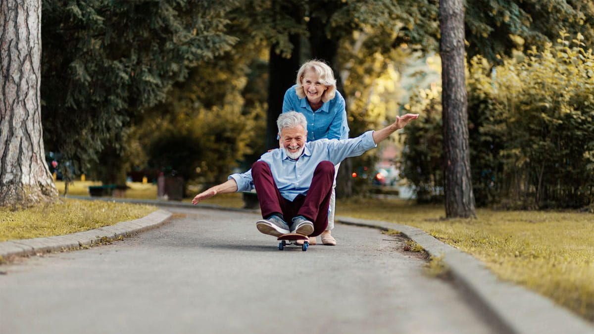 Ältere Frau schiebt ihren Mann auf einem Skateboard sitzend, lachend die Straße entlang