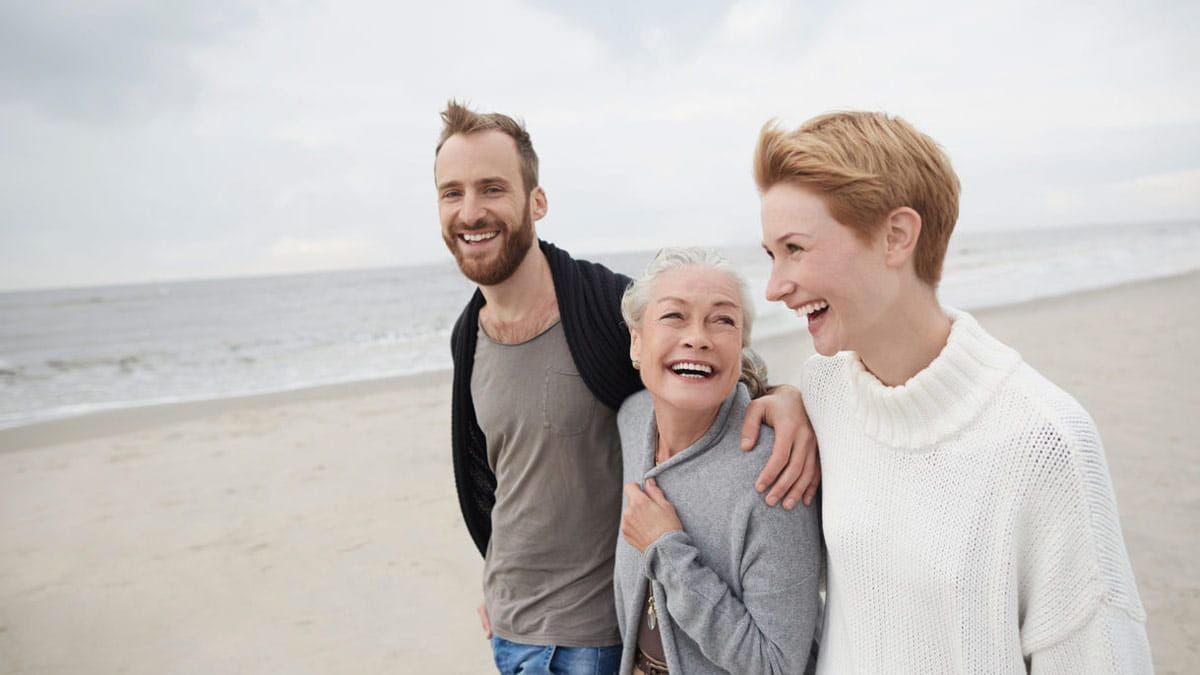 Ein Paar geht mit seiner Mutter lachend und Arm in Arm am Strand spazieren