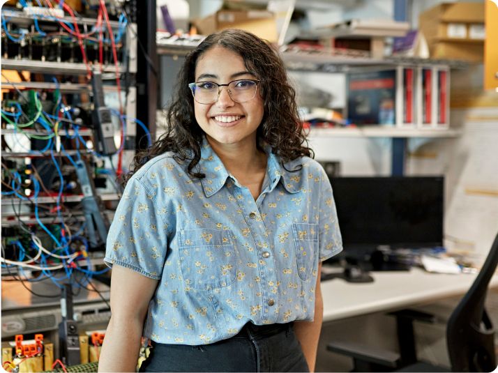 Junge Frau mit Brille lächelt in die Kamera und steht in einem Elektronik Labor.