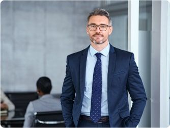 Geschäftsmann mit Brille und Anzug steht in einem Büro und blickt lächelnd in die Kamera.